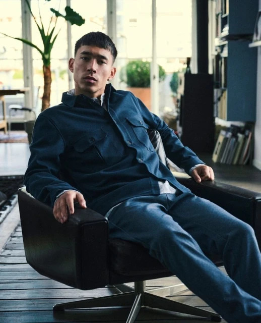A young adult male with short dark hair poses in a navy shirt and jeans, sitting in a black leather chair in a light-filled room.