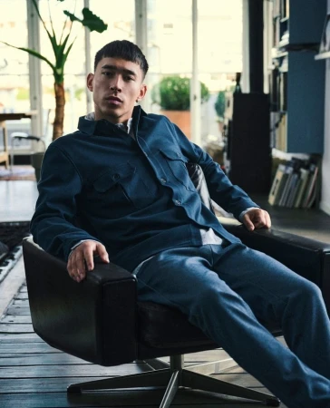 A young adult male with short dark hair poses in a navy shirt and jeans, sitting in a black leather chair in a light-filled room.