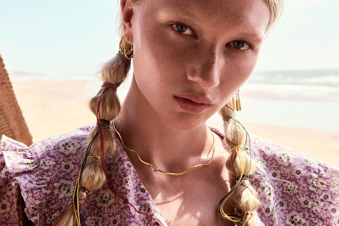 Close-up of an adult female model with fair skin, blonde braided hair adorned with strings, wearing a floral print dress and jewelry.