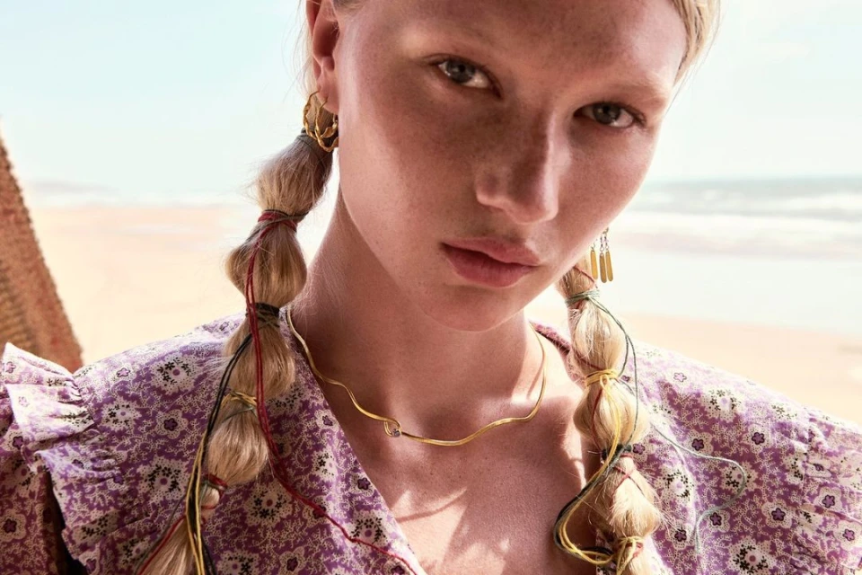 Close-up of an adult female model with fair skin, blonde braided hair adorned with strings, wearing a floral print dress and jewelry.