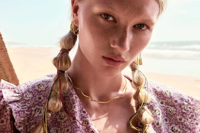 Close-up of an adult female model with fair skin, blonde braided hair adorned with strings, wearing a floral print dress and jewelry.