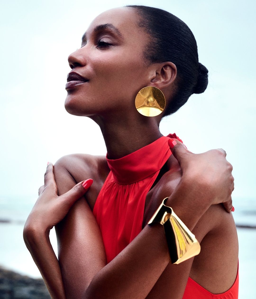 Dark-skinned woman in a red dress with gold jewelry poses outdoors against a blurry light-blue and white background. Fashion shot.