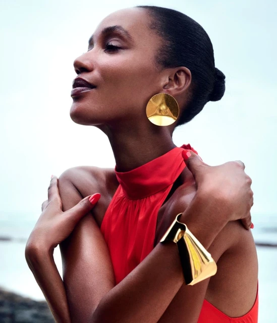 Dark-skinned woman in a red dress with gold jewelry poses outdoors against a blurry light-blue and white background. Fashion shot.