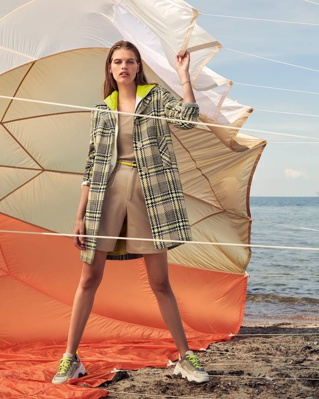A fashion model poses on a beach wearing a plaid coat, neutral shorts, tank top, and sneakers with a parachute in the background.