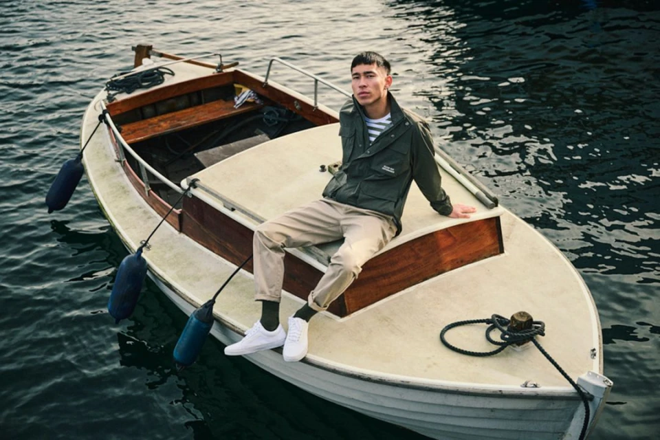 Young adult male sits on a white rowboat in water, wearing an olive jacket, beige pants, and white sneakers, in a lifestyle fashion shot.