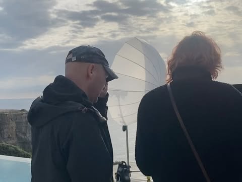 Two adults outdoors on an overcast day. One is bald and wearing a cap and dark jacket, while the other has auburn hair and a black coat.