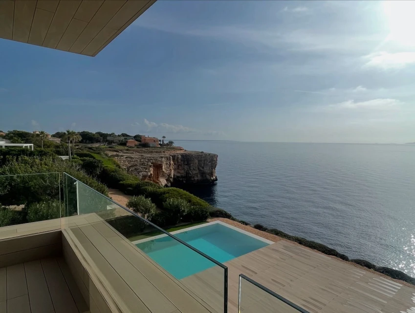 Coastal view featuring a modern terrace with clear glass railings, turquoise pool, and ocean cliffs under a partly cloudy sky.
