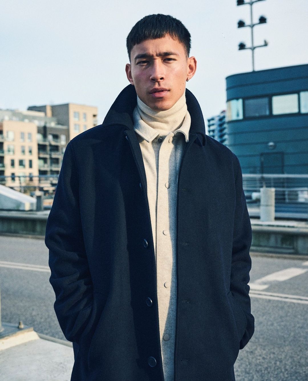 A young adult man stands on a city street wearing a dark overcoat, cream turtleneck, and light gray shirt with buildings in the background.