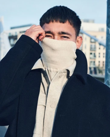Close-up portrait of a man wearing a dark coat and cream turtleneck covering his face, photographed in London.