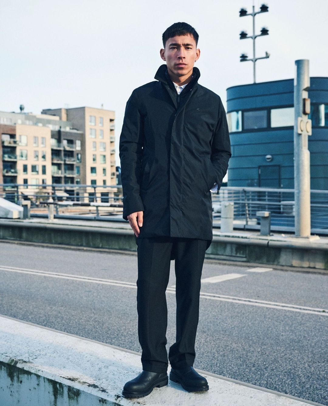 An Asian man in a black coat stands on a bridge with urban buildings, a utility pole, and an overcast sky in the background.