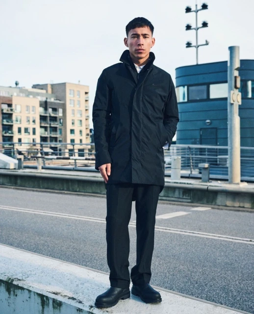 An Asian man in a black coat stands on a bridge with urban buildings, a utility pole, and an overcast sky in the background.