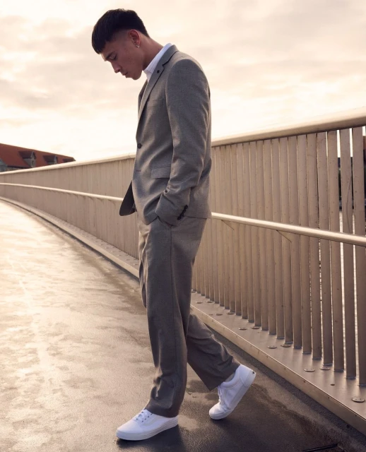 Adult male in a grey suit and white sneakers walks along a modern pedestrian bridge under a cloudy sky, showcasing urban fashion.