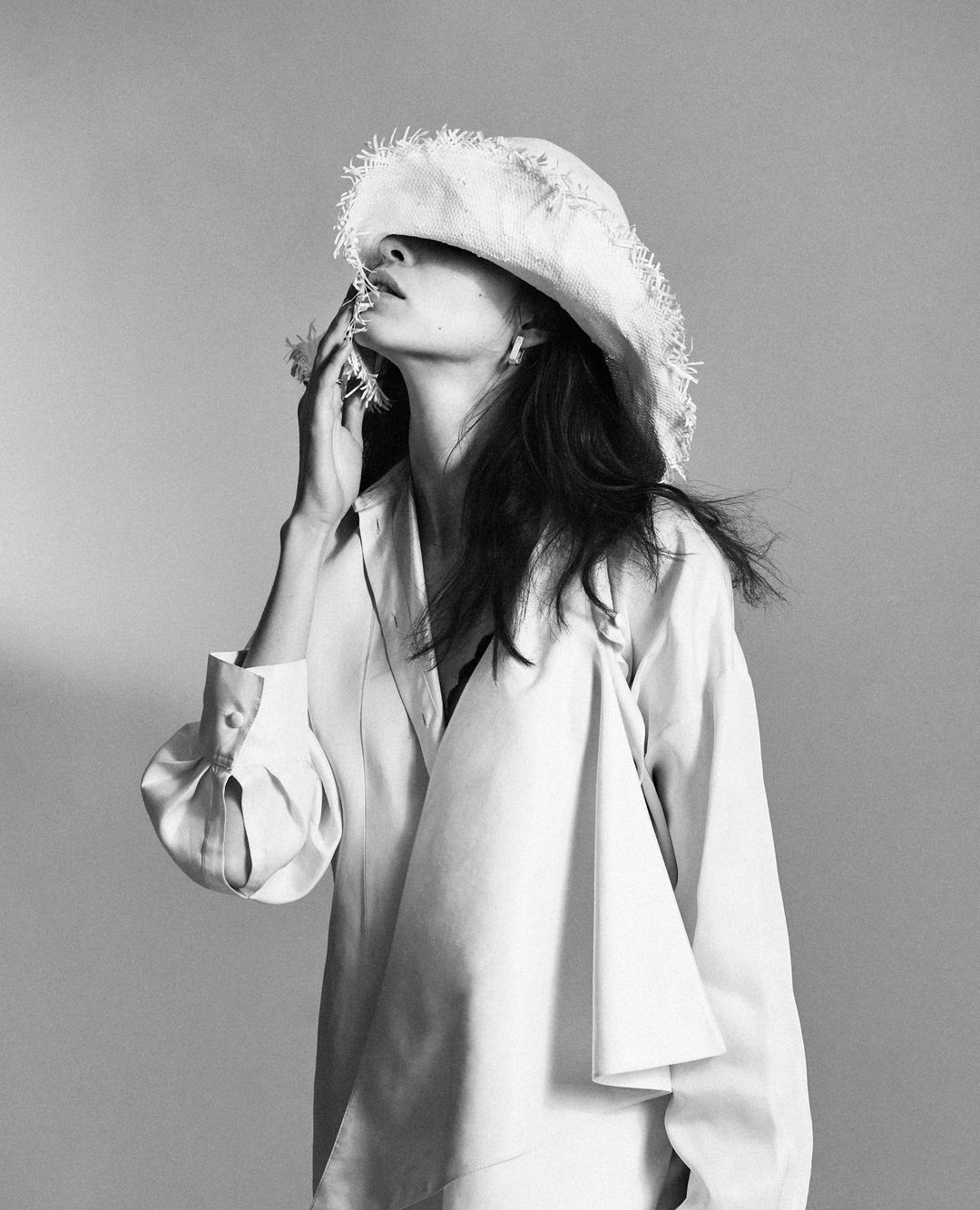 Monochrome portrait of a woman wearing an oversized shirt and a fringed hat, posing with her hand near her face in a studio setting.