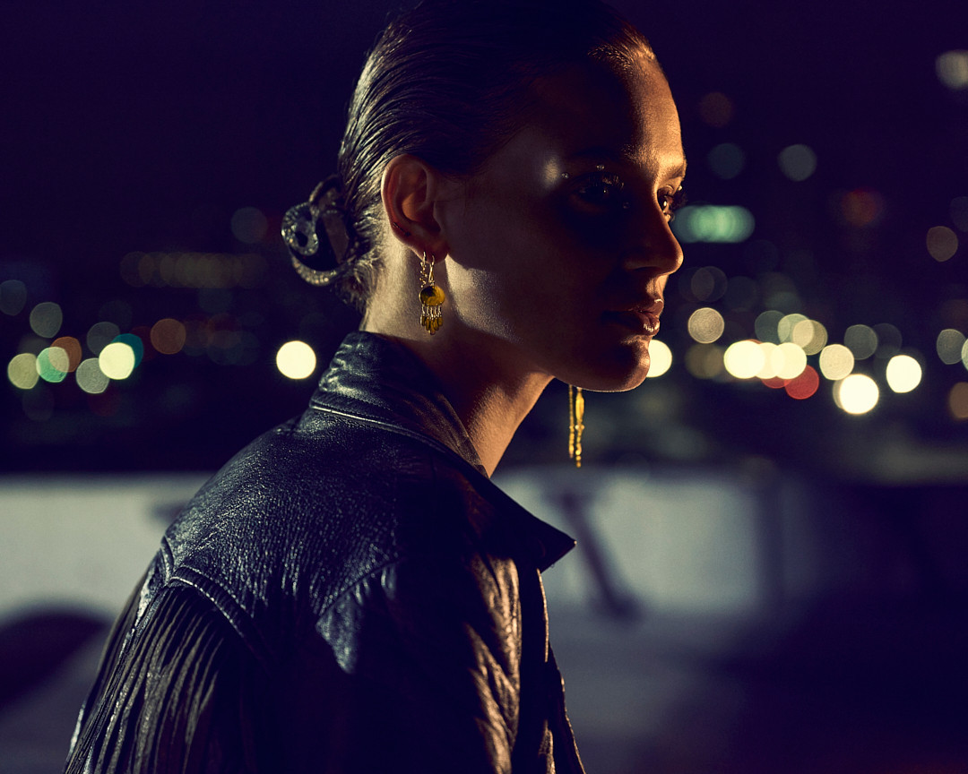 Woman in leather jacket poses at night with city lights. She wears drop earrings and has hair slicked back in dim, cinematic lighting.