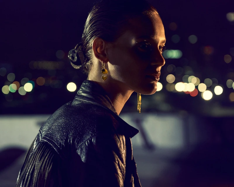 Woman in leather jacket poses at night with city lights. She wears drop earrings and has hair slicked back in dim, cinematic lighting.