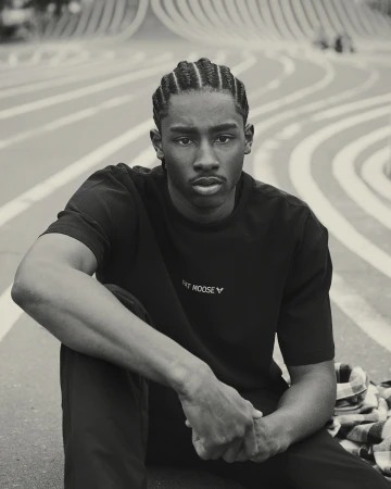 Portrait of a young Black man with braided hair, wearing a black t-shirt, sitting outdoors with striped track lines in the background.