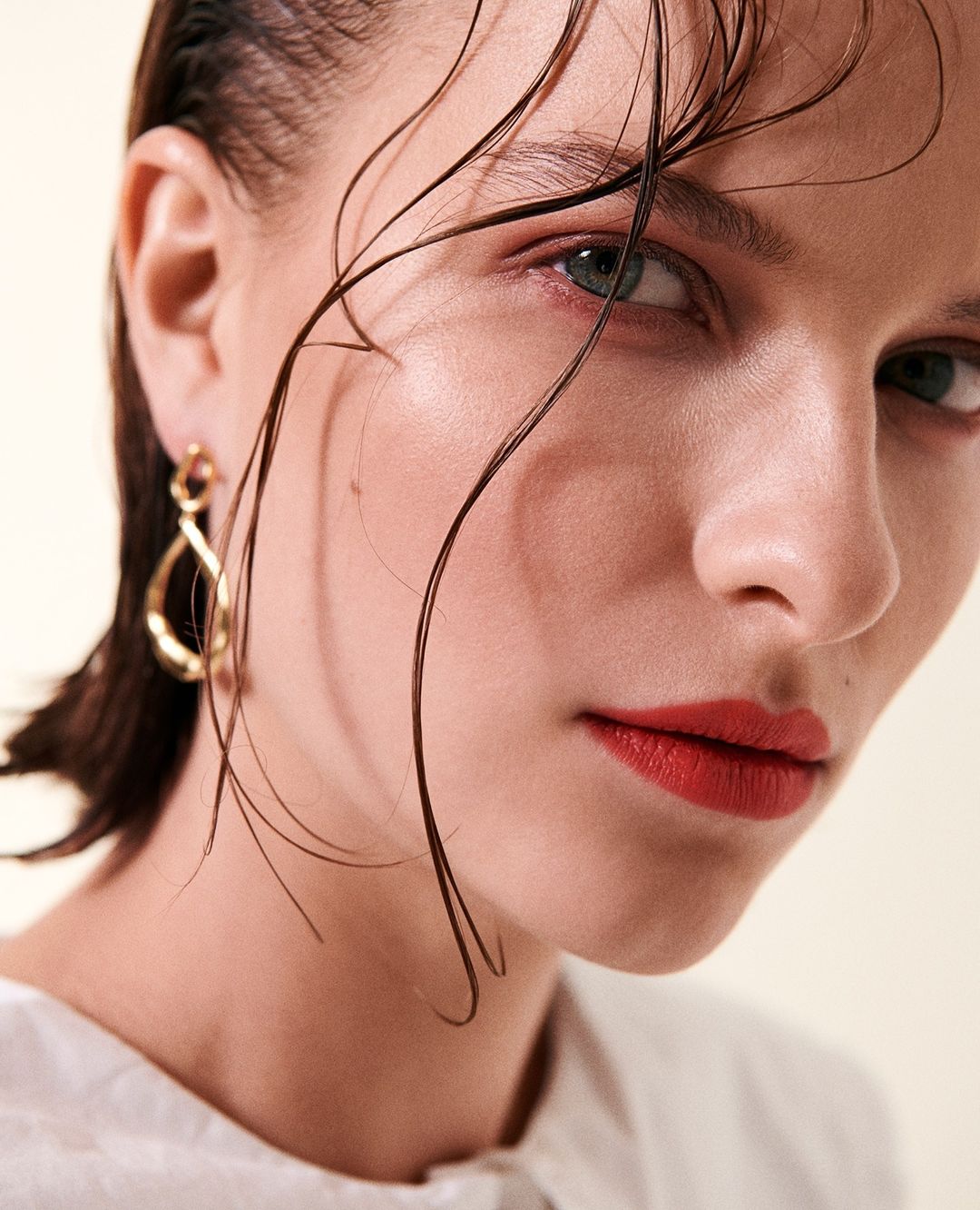 Close-up portrait of a young fair-skinned woman with wet hair and bright red lipstick wearing gold earrings and a white top.