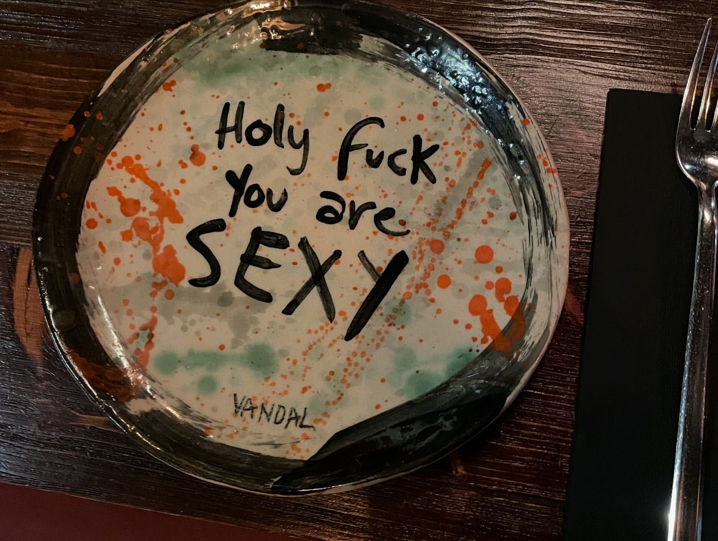 A handmade ceramic plate with an expressive inscription sits on a wooden table next to silverware, creating an artistic dining scene.