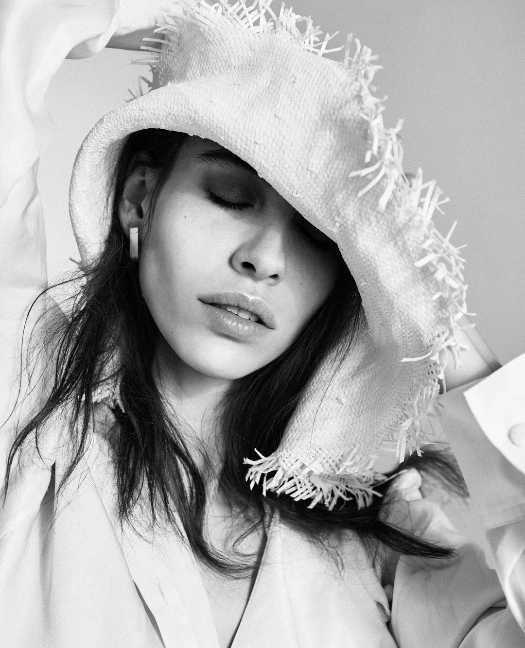 Monochrome portrait of a woman wearing a textured hat and an unbuttoned shirt, eyes closed and face softly illuminated by natural light.