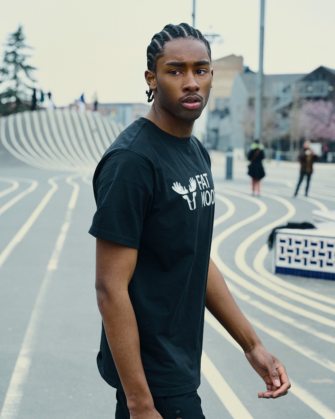 Man in black t-shirt stands outdoors in an urban setting with striped ground, captured in a medium shot with a blurred background.