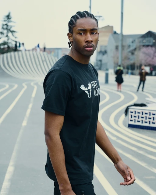 Man in black t-shirt stands outdoors in an urban setting with striped ground, captured in a medium shot with a blurred background.