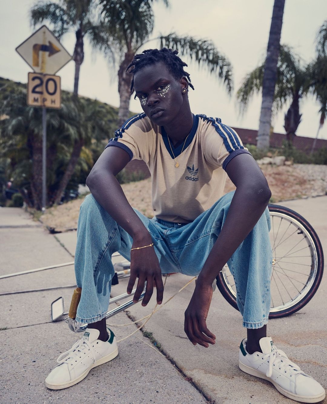 Young Black man with glitter makeup poses on bicycle wearing Adidas top, light blue jeans, and white sneakers in an urban outdoor setting.