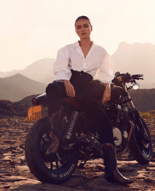 A woman sits on a motorcycle on a rocky plain with mountains in the background, wearing a white shirt and cowboy boots.