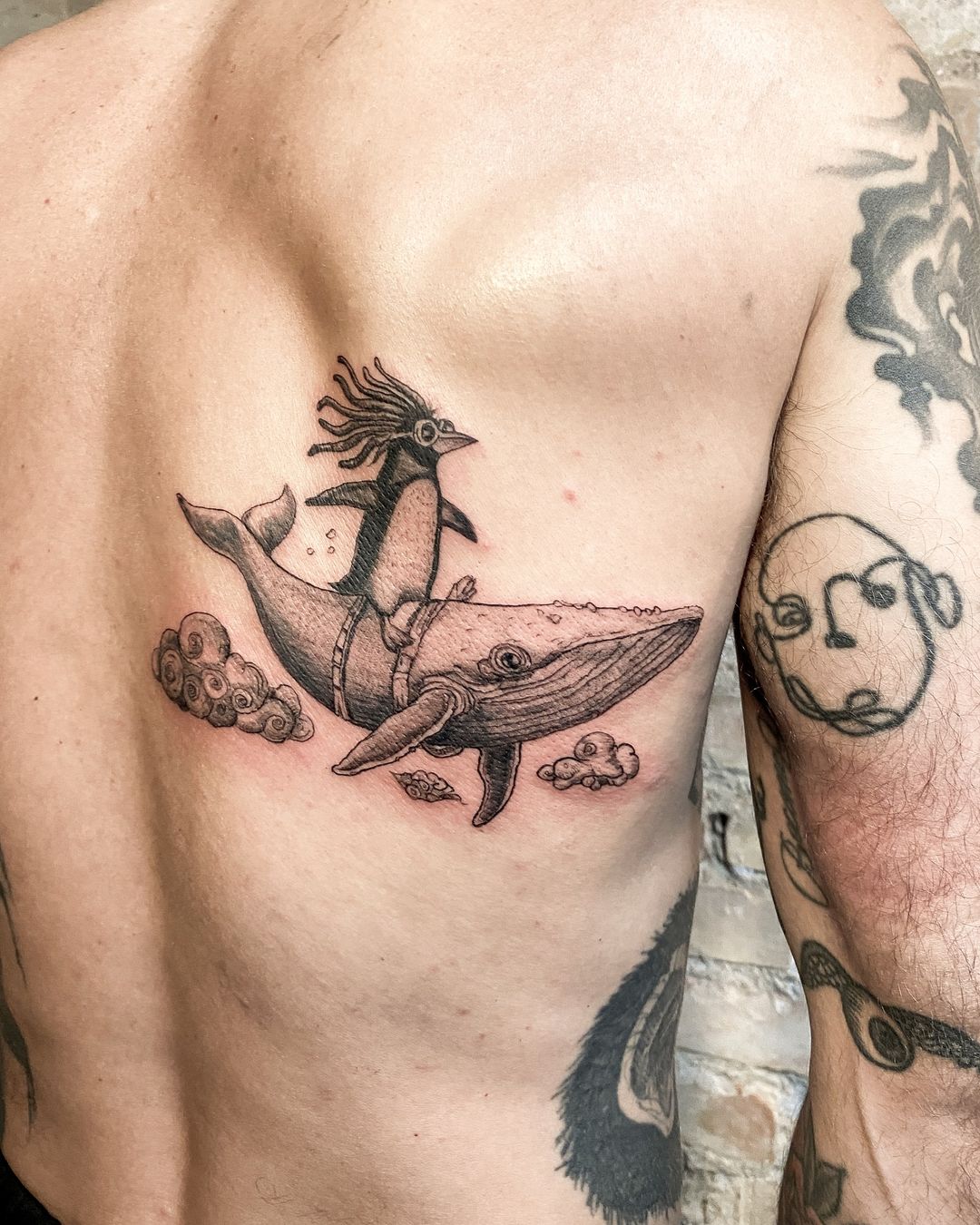 Back tattoo of a penguin with dreadlocks riding a whale, surrounded by clouds, done in black and gray ink on a human's torso.