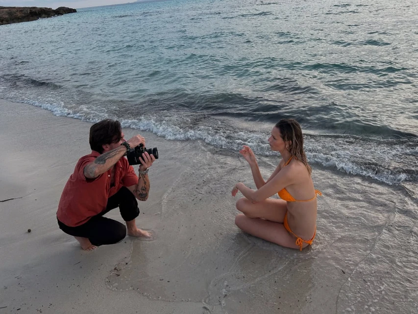 A photographer captures a woman in an orange bikini, seated in the shallow water of a beach with an ocean and rocky coast backdrop.