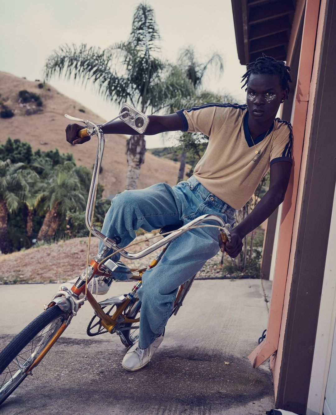 Dark-skinned adult wearing blue jeans rides an old-fashioned bicycle in an outdoor setting on a sunny day; palm trees in the background.