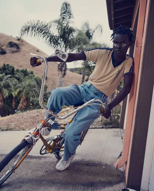 Dark-skinned adult wearing blue jeans rides an old-fashioned bicycle in an outdoor setting on a sunny day; palm trees in the background.