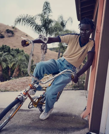 Dark-skinned adult wearing blue jeans rides an old-fashioned bicycle in an outdoor setting on a sunny day; palm trees in the background.