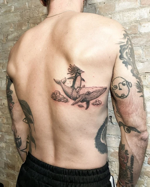 Tattoo on a man's back depicting a whale with a crested penguin rider above some clouds. The tattoo is rendered in shades of grey.