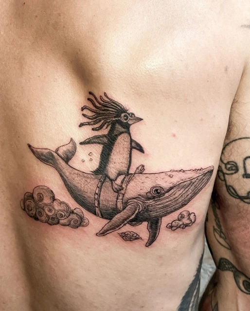 Tattoo art on skin: A penguin with dreadlocks rides a whale amidst cloud-like formations, showcasing intricate black and grey ink detailing.