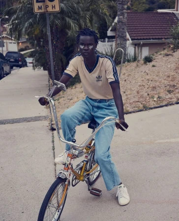 Young adult on a vintage-style bicycle with high handlebars in a suburban neighborhood, wearing a t-shirt and jeans.