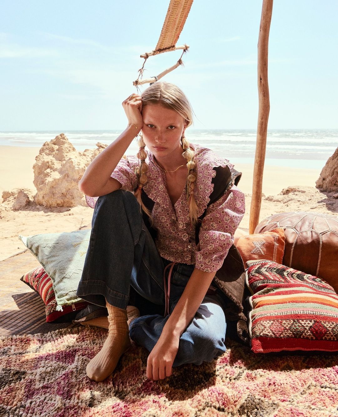 Woman in floral shirt and denim pants sitting on patterned rug near beach with ocean in background on sunny day.
