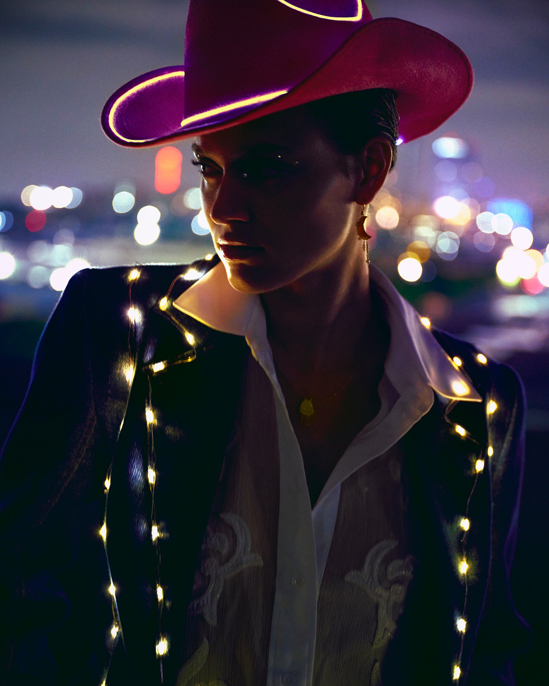 Glamorous woman in a cowboy hat adorned with fairy lights poses in front of a blurred city skyline at dusk.