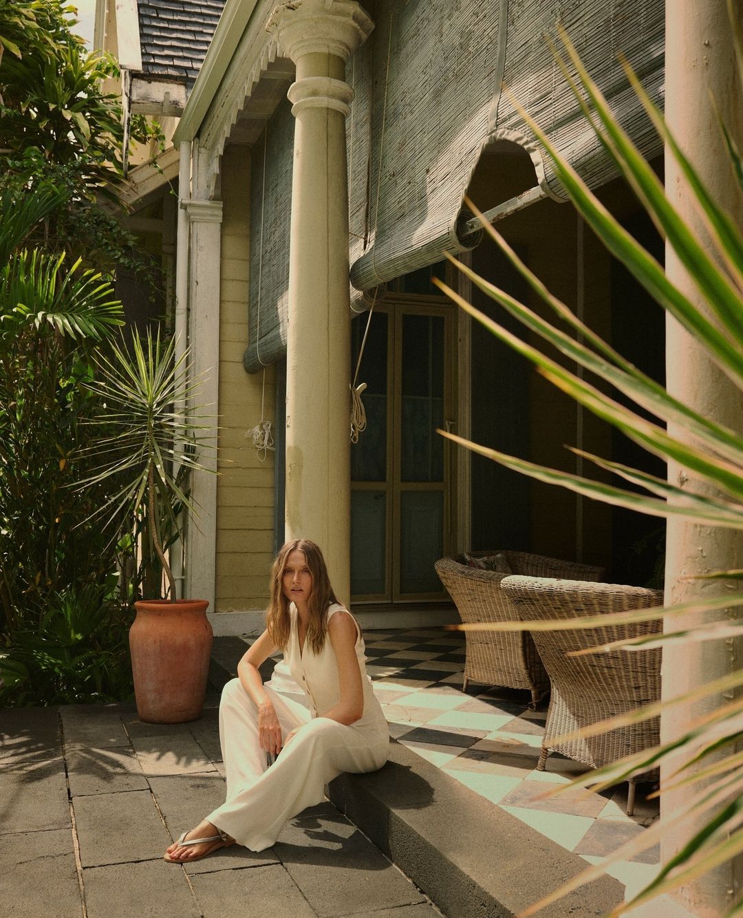 Woman in white jumpsuit sits outside on a porch with pillars and wicker chairs, surrounded by greenery and a neutral color palette.