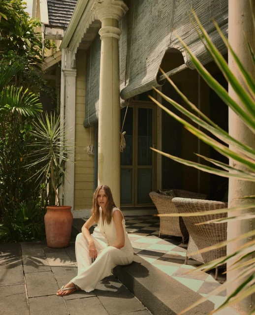 Woman in white jumpsuit sits outside on a porch with pillars and wicker chairs, surrounded by greenery and a neutral color palette.