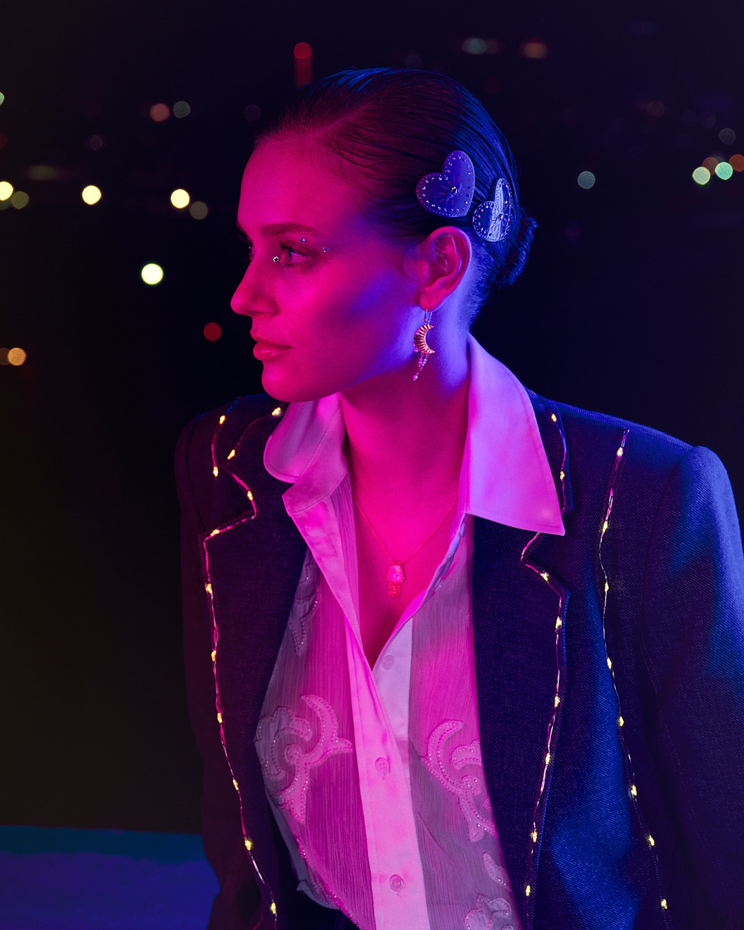 Side profile of a young woman with heart-shaped hair clips, moon earrings, and a jacket with light string details against a bokeh background.