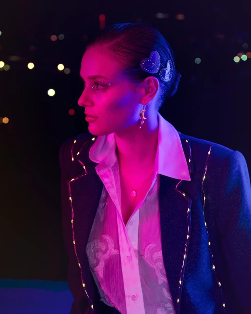 Side profile of a young woman with heart-shaped hair clips, moon earrings, and a jacket with light string details against a bokeh background.