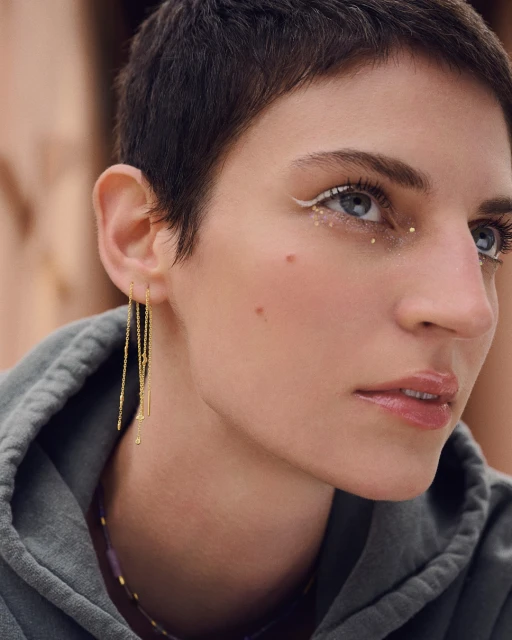 Close up of a woman with a pixie cut wearing gold earrings and white eyeliner, viewed from the side with soft, diffused lighting.