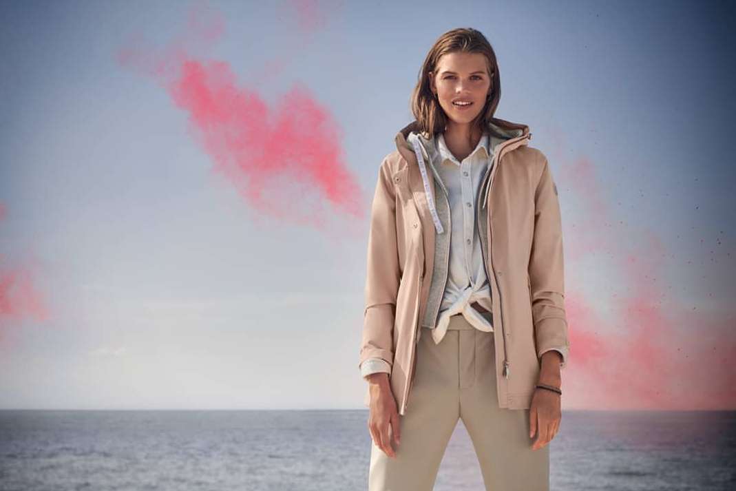 Woman in a tan hooded jacket and pants standing outside with a seascape and pink clouds in the background, fashion advertising shot.