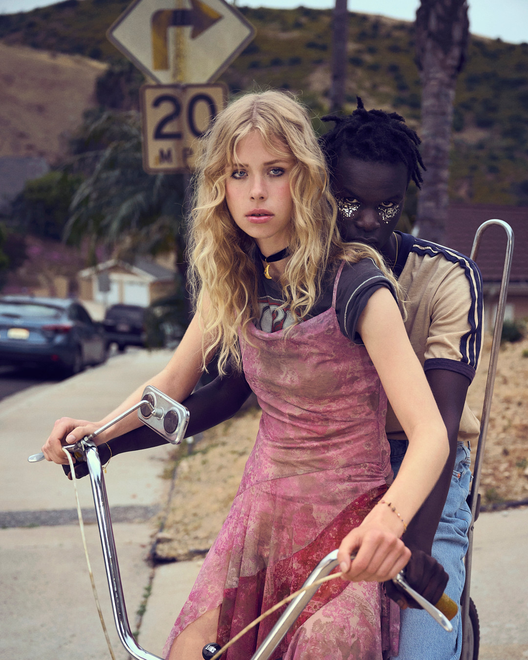Two models on a bike under a speed limit sign. One blonde model wears a pink dress, and the other model has embellished makeup.