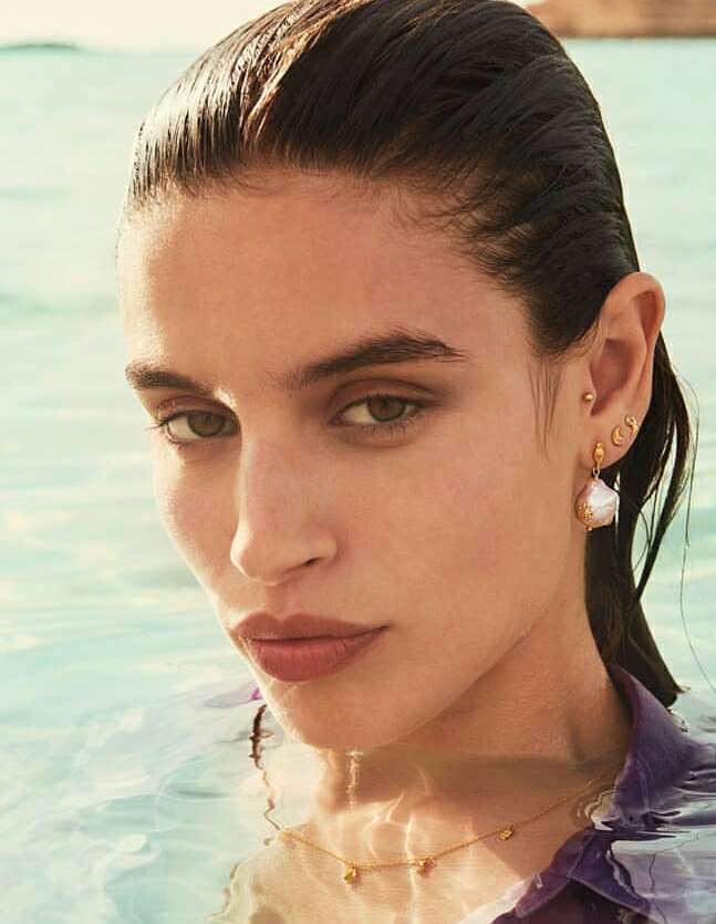 Close-up of a woman in water wearing gold jewelry, including a necklace and earrings, with natural light and a serene expression.