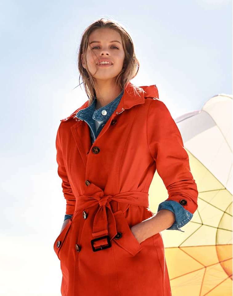 A woman in a red trench coat stands outside on a sunny day with her hands in her pockets, looking towards the camera with a smile.