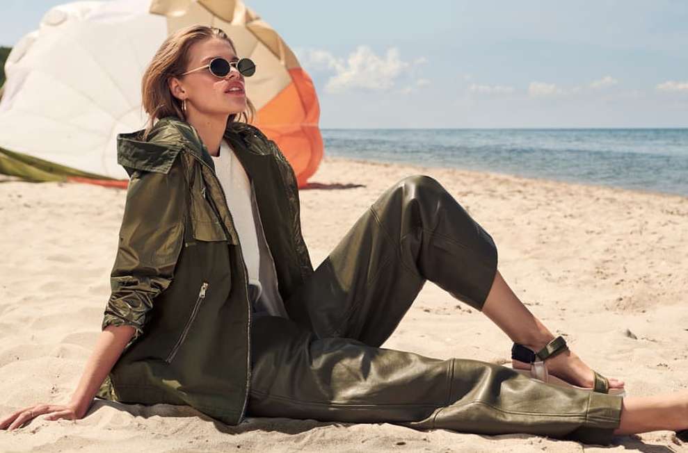 Adult female model sits on a sandy beach near the ocean wearing a white shirt, green jacket, green pants, and sandals. A tent is visible.