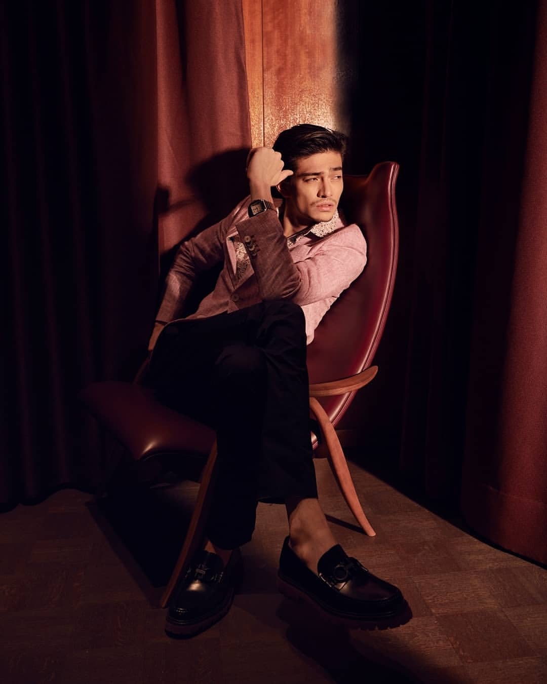 Portrait of a young adult male model sitting in a red chair wearing a blazer, trousers, and loafers, against a dark red curtain backdrop.