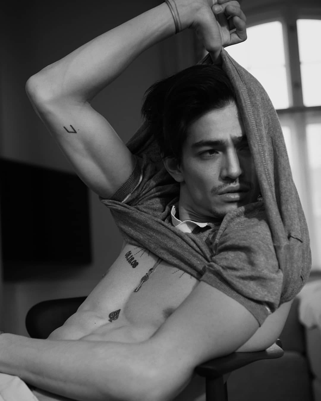 Black and white portrait of a shirtless man with tattoos, pulling a gray sweater over his head in an indoor setting with window light.