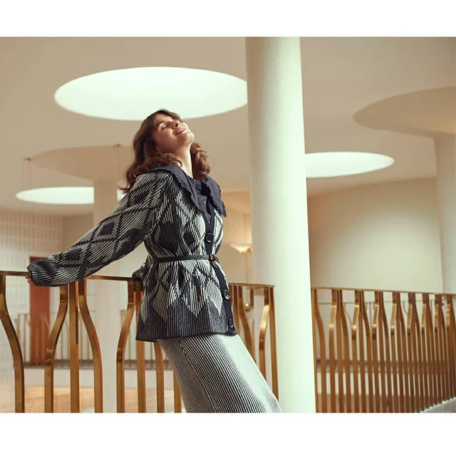 Woman in patterned cardigan and striped skirt poses indoors with head tilted back and eyes closed, near decorative railing and white pillar.